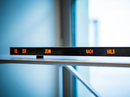 Längliche Leuchtschrift-Uhr auf einem weissen Regal vor einem Fenster. Der Satzteil "ES IST ZEHN NACH HALB" sind in orangefarbener Schrift auf der dunklen Frontfläche beleuchtet, während weitere Buchstaben dezent im Hintergrund sichtbar bleiben. Die Uhr ist horizontal ausgerichtet und modern gestaltet, das Tageslicht fällt weich durch das Fenster im Hintergrund.