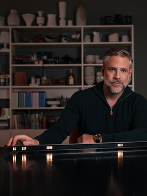 Michael Walter sitzt an einem Tisch und hält eine längliche Leuchtschrift-Uhr mit den angezeigten Satz "ES IST ZEHN NACH". Die Uhr steht auf einer glänzenden Oberfläche und leuchtet in warmem Licht. Im Hintergrund befindet sich ein Regal mit Büchern, Keramik und Dekorationsgegenständen. Die Umgebung ist wohnlich und stimmungsvoll beleuchtet.