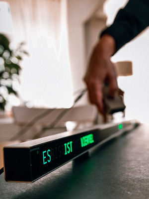 Längliche Leuchtschrift-Uhr auf einer dunklen Oberfläche in einem hellen Raum mit Wohnatmosphäre. Der Satzteil "ES IST VIERTEL" leuchten in grün auf der Frontfläche. Eine Hand im Hintergrund hält ein Bauteil, während weiches Tageslicht durch Vorhänge einfällt. Die Szene zeigt eine Kombination aus Technik und wohnlichem Umfeld.
