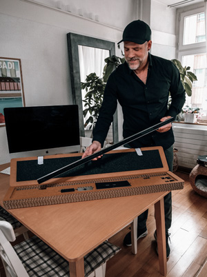 Michael Walter steht in einem Wohnraum an einem Holztisch und hebt eine längliche Leuchtschrift-Uhr aus einer geöffneten Verpackung. Die Verpackung aus Wellkarton enthält Schaumstoffeinlagen und geordnetes Zubehör. Im Hintergrund befinden sich ein Computer, ein Spiegel, Zimmerpflanzen und ein Fenster mit Tageslicht. Die Szene wirkt wohnlich und zeigt den Moment des Auspackens.
