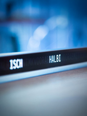 Längliche Leuchtschrift-Uhr auf einer hellen Oberfläche mit den Wörtern ISCH und HALBI in weisser Schrift beleuchtet. Die restlichen Buchstaben sind schwach im Hintergrund erkennbar. Die Umgebung ist von kühlem, blauem Licht geprägt, im unscharfen Hintergrund sind Fenster oder Glasflächen sichtbar. Die Szene wirkt ruhig und modern.
