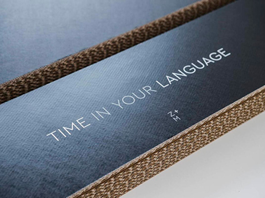 Nahaufnahme einer Verpackung aus brauner Wellpappe mit schwarzer, leicht strukturierter Oberfläche. Auf der Oberseite ist in weisser Schrift der Text TIME IN YOUR LANGUAGE sowie das Z+M-Logo aufgedruckt. Die Kante zeigt die wellige Struktur des Kartonmaterials. Die Verpackung wirkt stabil und hochwertig verarbeitet.